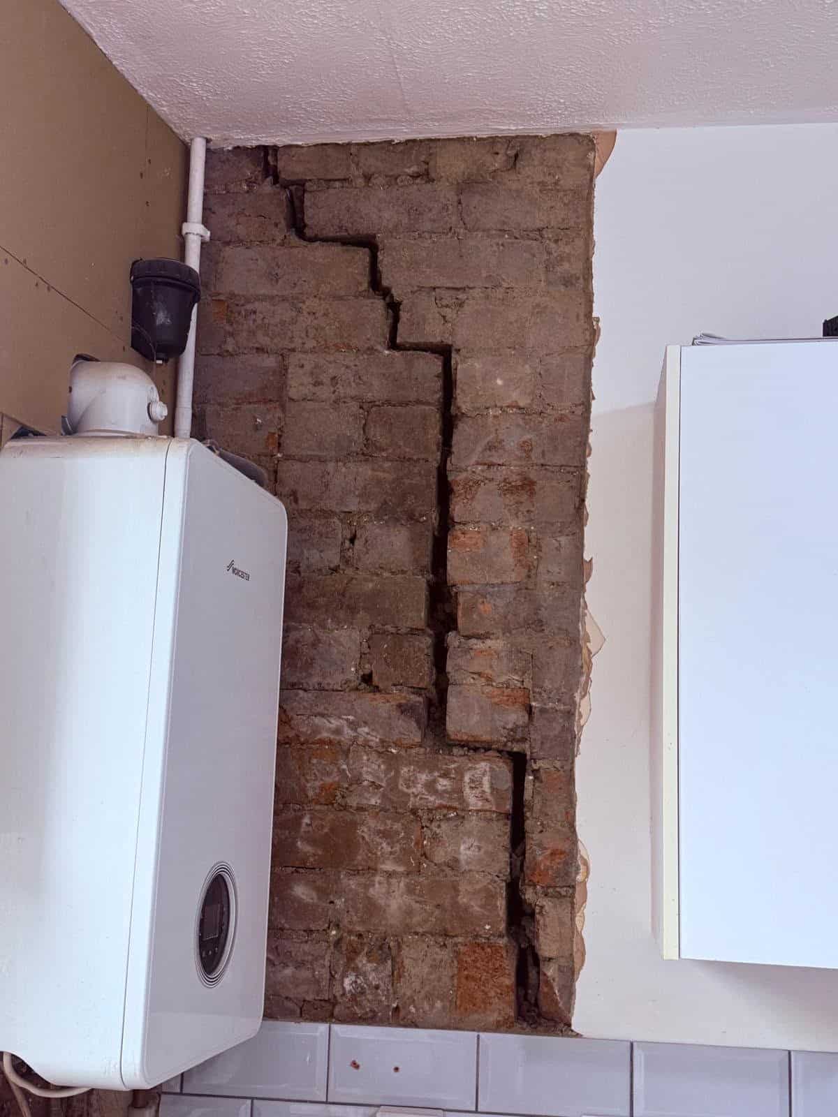 Kitchen wall backing onto the chimney breast stripped to bare brick, showing deteriorated masonry behind the boiler