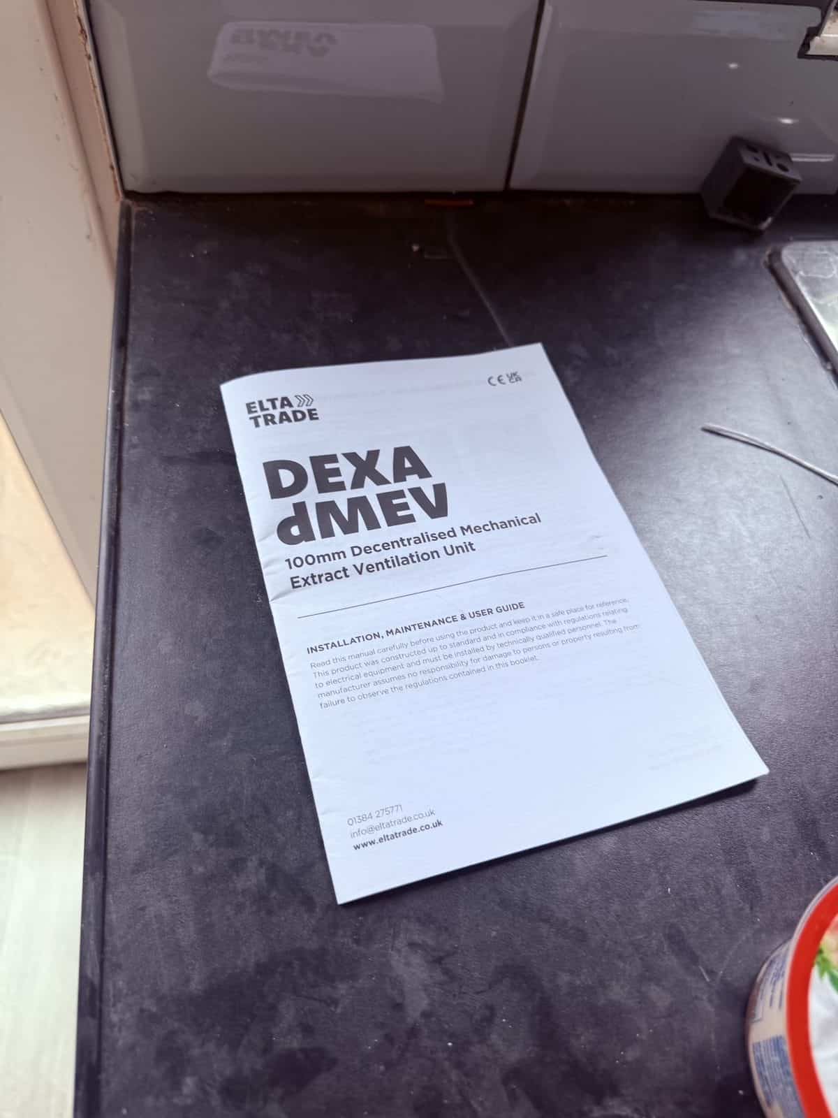 Elta DEXA dMEV 100 installation manual open on a kitchen worktop during the fan install