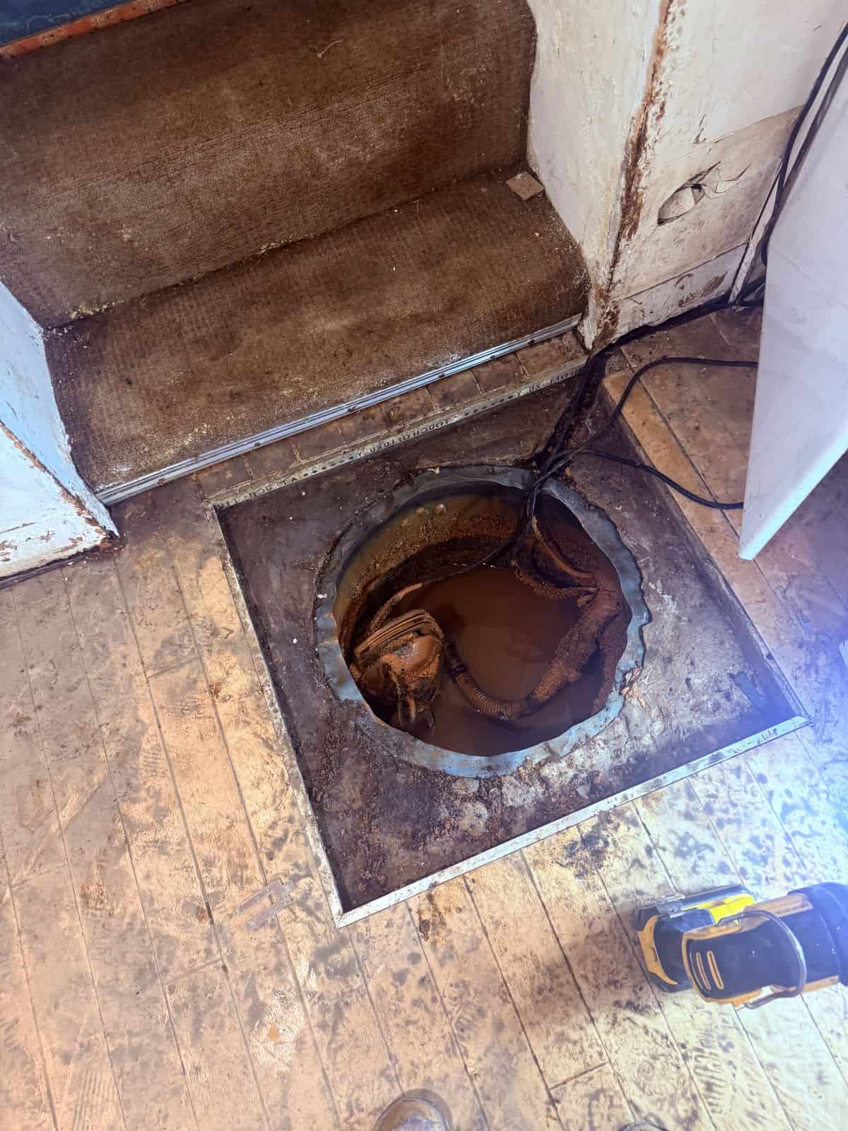 Corroded sump chamber cut into a timber cellar floor at the foot of the cellar steps